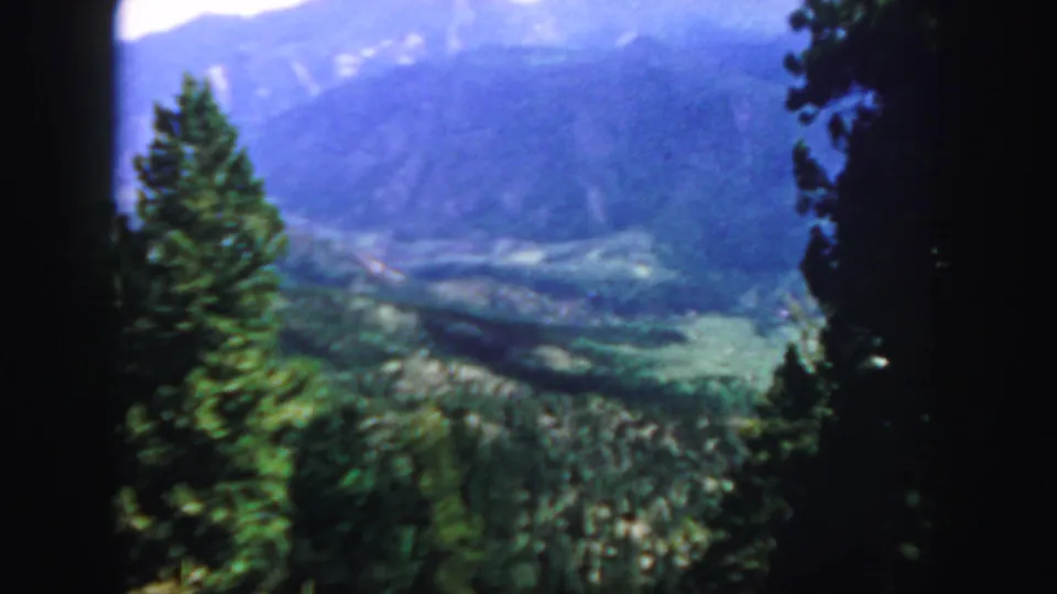 1958 Colorado 8mm frame showing an evergreen foreground and a broad green valley below distant mountain ridges.