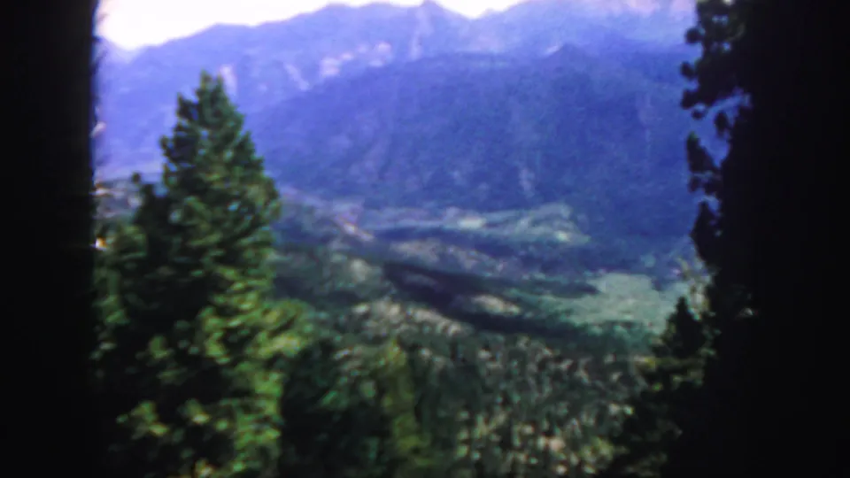 1958 Colorado home-movie frame showing a central evergreen, forested valley, and hazy mountain ridges beyond.