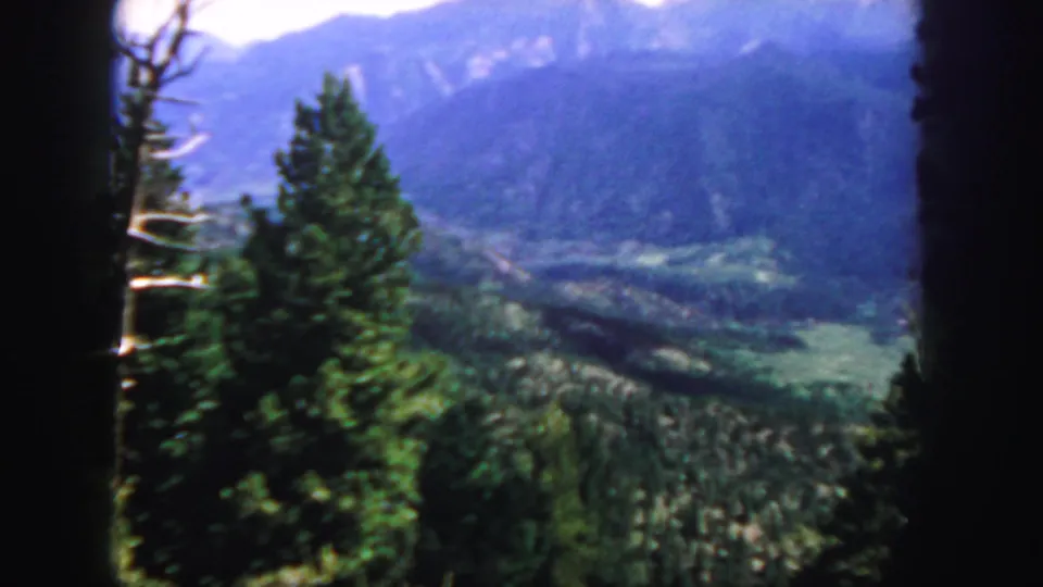 1958 Colorado 8mm frame with foreground conifers, a wide green valley, and hazy blue mountain slopes in the distance.