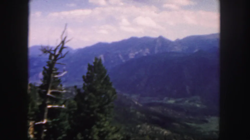 1958 Colorado scenic frame with a green valley below blue mountains, softened by 8mm focus and film grain.