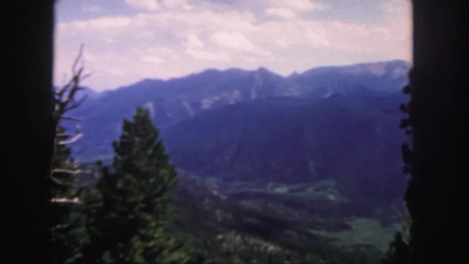 Colorado 8mm home-movie frame showing a mountain panorama, foreground evergreens, a bare tree, and valley landscape.
