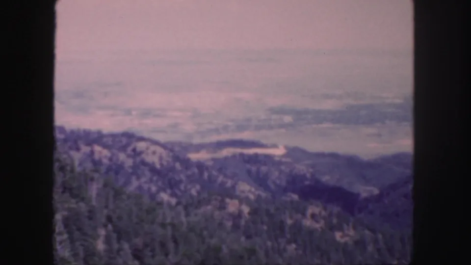 1958 Colorado 8mm frame showing evergreen-covered hills, distant valley landforms, and a muted hazy sky.