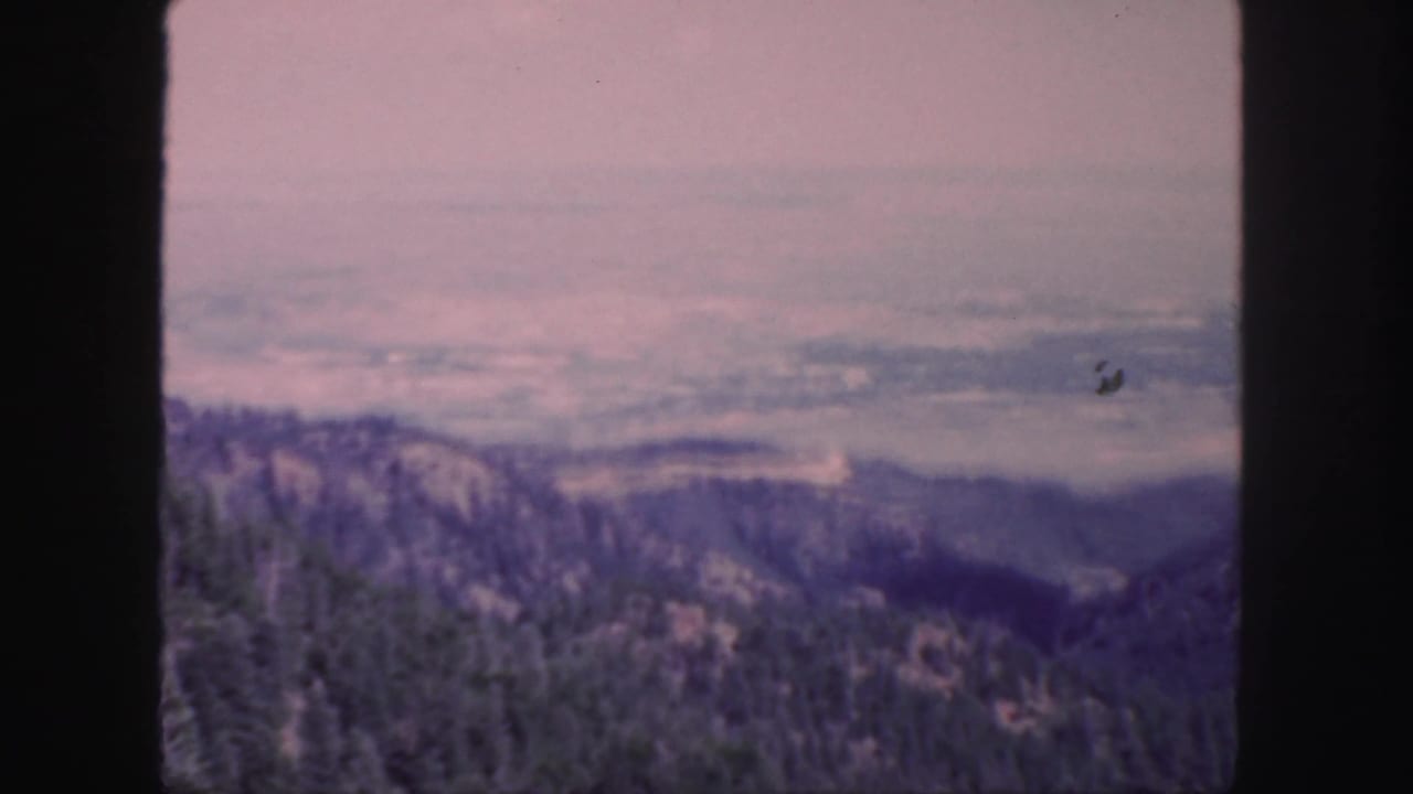 1958 Colorado 8mm frame showing forested hills, distant ridgelines, pale haze, and dark film gate borders.