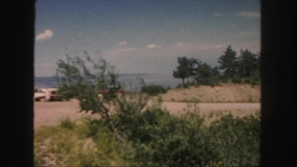 1958 Colorado roadside view with brush, a sandy rise, trees, pale sky, and a small light-colored vehicle in the distance.