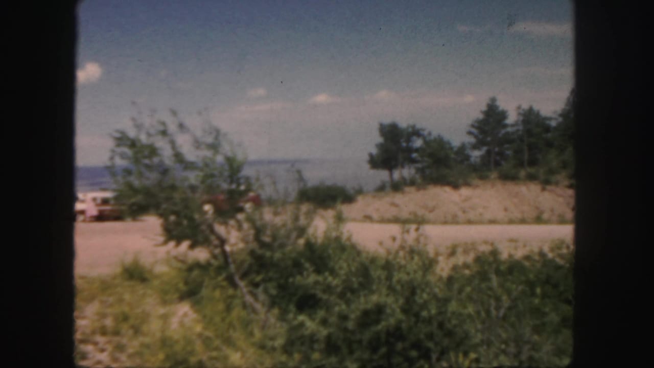 Roadside shrubs and low hills seen through a dark vehicle window frame.