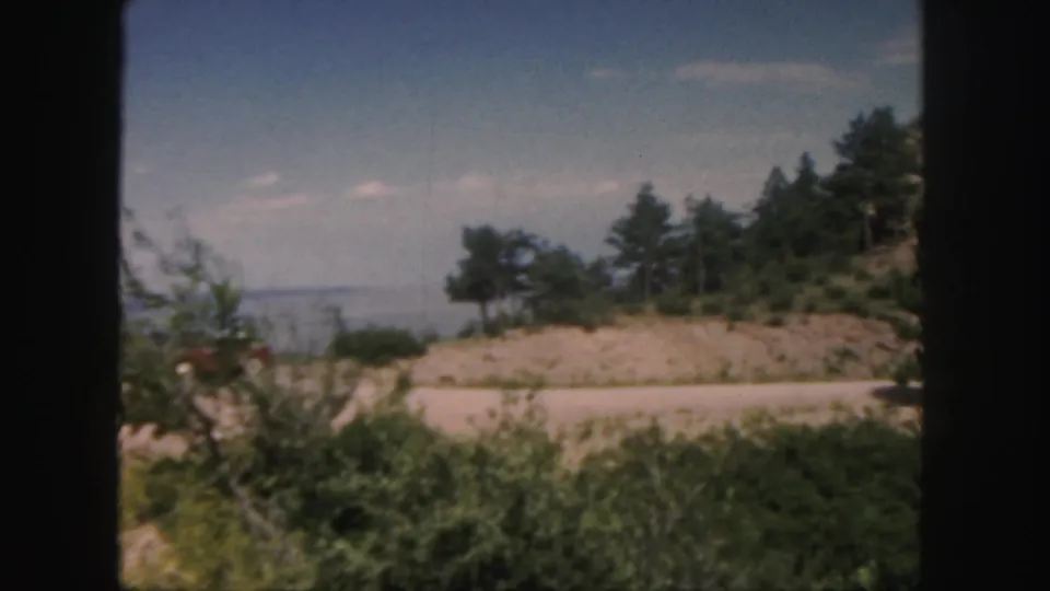 Colorado home-movie view from a moving vehicle, showing roadside brush, a low tree-covered hill, distant haze, and blue sky.