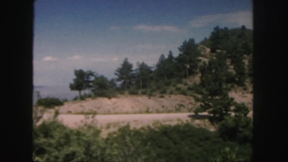 1958 Colorado landscape frame with roadside vegetation, a pale road or shoulder, green hillside trees, and soft blue sky from a moving car.