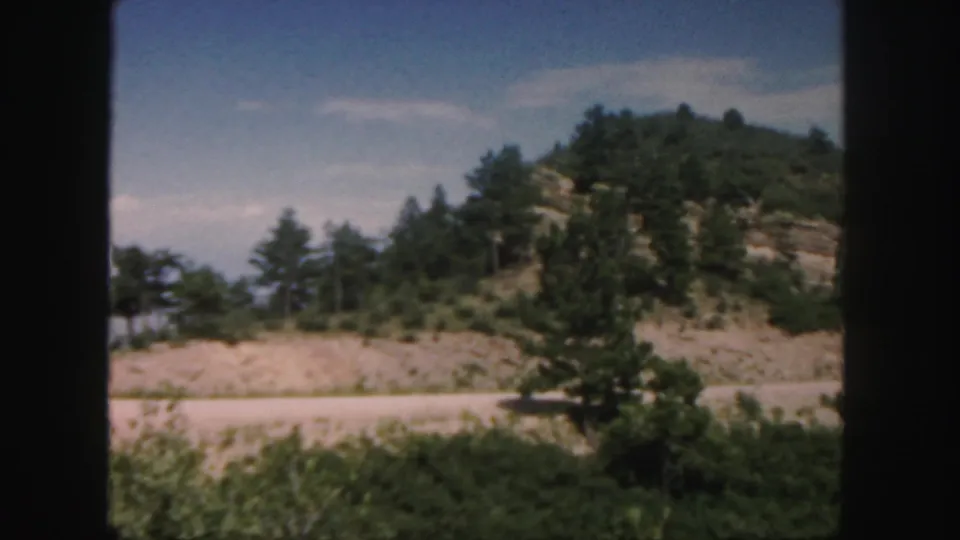 Colorado 8mm road-trip view of shrubs, a pale roadside strip, and a rocky hill with scattered trees beneath a blue sky.