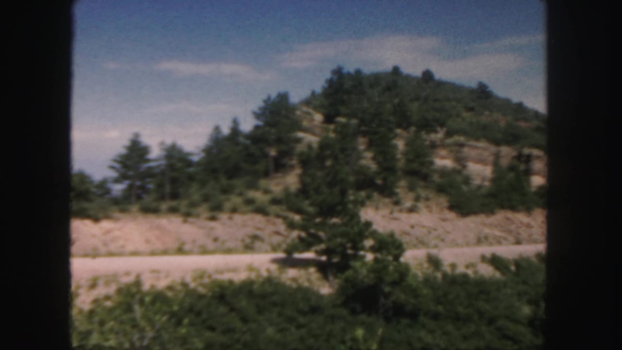 Rocky hillside with pine trees above a pale road, framed by dark vertical edges.