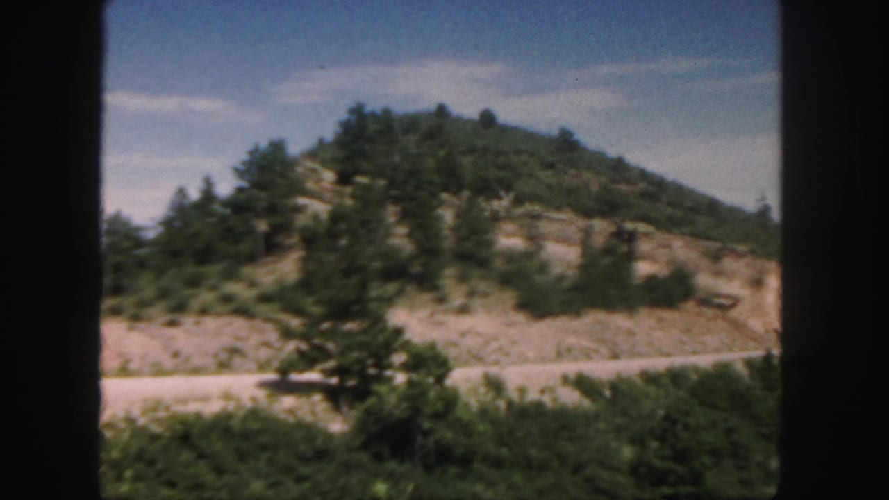 1958 Colorado roadside view with pine trees, exposed rock formations, brush, and a pale road.