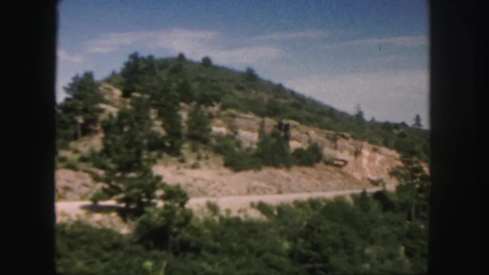 1958 Colorado 8mm frame showing a roadside hill with scattered pines and reddish exposed rock.