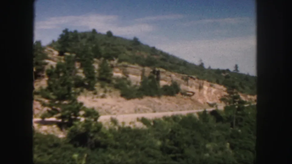 1958 Colorado road footage showing a pine-dotted rocky hillside, roadside brush, and blue sky.