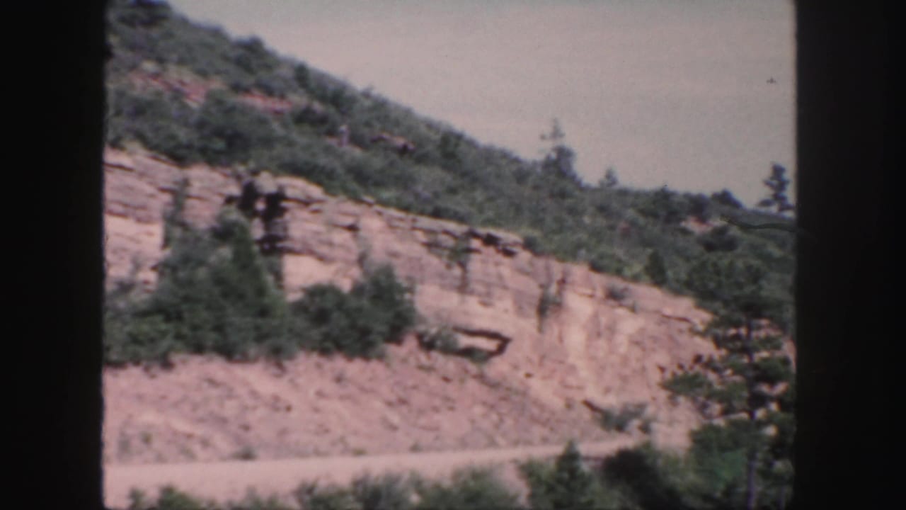 1958 Colorado 8mm frame showing an exposed rocky slope, brush, and a dirt road at the base with no people or vehicles visible.