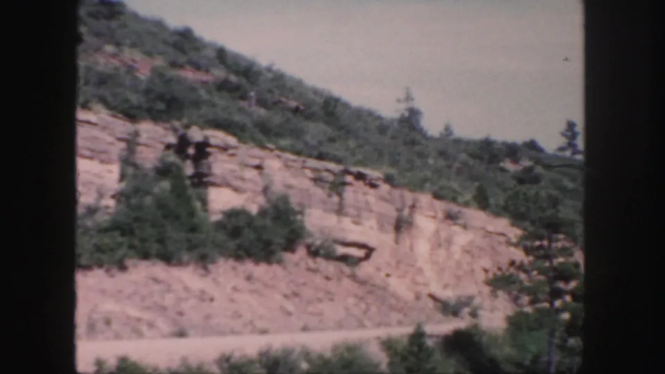 1958 Colorado 8mm frame showing an exposed rocky slope, brush, and a dirt road at the base with no people or vehicles visible.