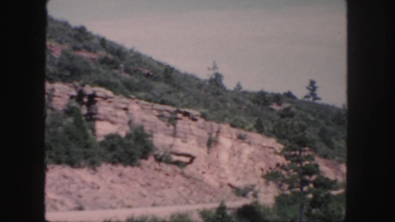 Mountain cut with exposed rock layers, trees, and road edge beneath a cloudy sky.
