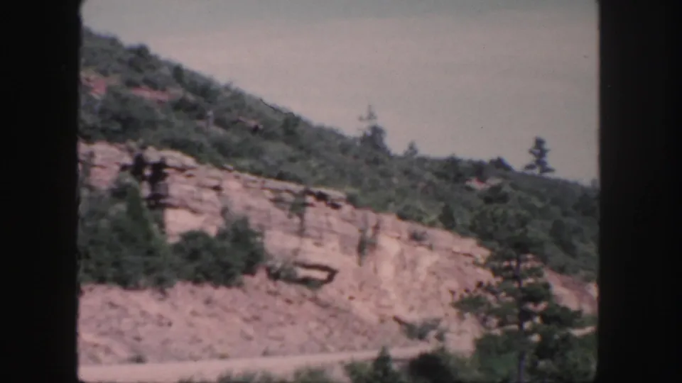 1958 Colorado roadside view of a steep rock face and brush above a dirt road, framed by dark 8mm film edges.