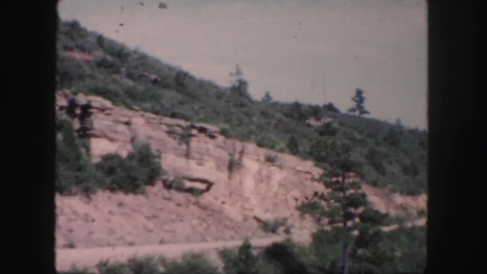Colorado home-movie frame from 1958 showing a steep roadside slope with reddish rock, vegetation, and no visible traffic.