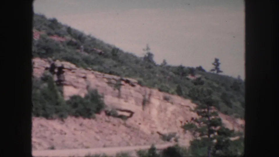 Late-1950s Colorado 8mm frame of a rural mountain road below exposed rock and trees, with no people, vehicles, or signs visible.