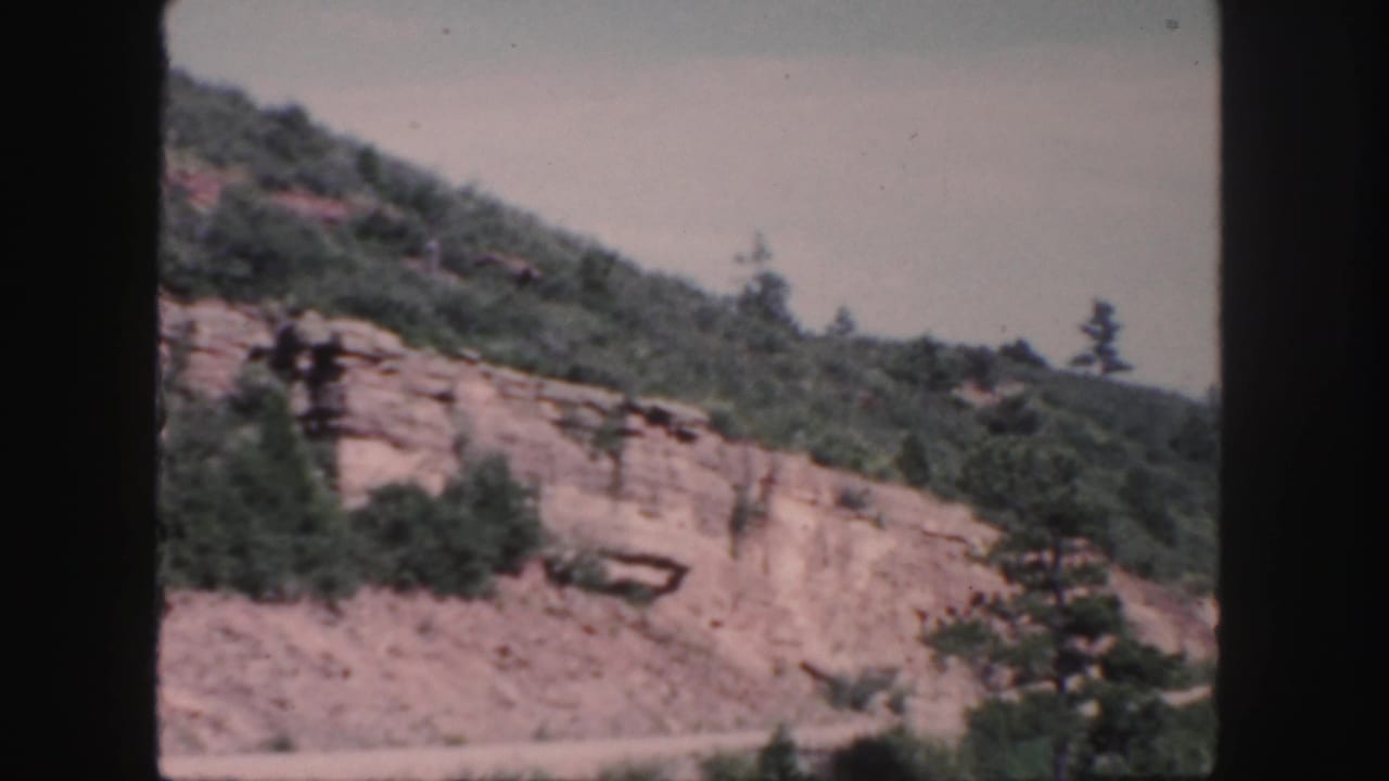 Colorado mountain roadside scenery on 8mm film, showing a rocky cut, scrub trees, and a soft pale sky.
