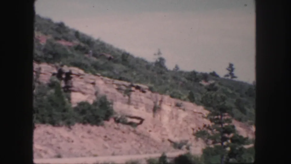 Colorado mountain roadside scenery on 8mm film, showing a rocky cut, scrub trees, and a soft pale sky.