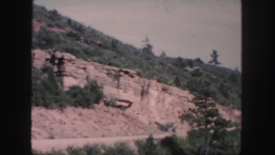 A Colorado mountain road cut appears in soft 8mm color, with trees along the ridge and pale exposed rock below.