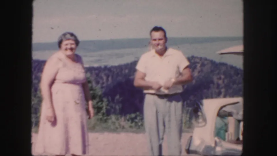In supported 1958 Colorado footage, a woman in a pink dress and a man in a light shirt stand near a classic car with mountain views behind them.