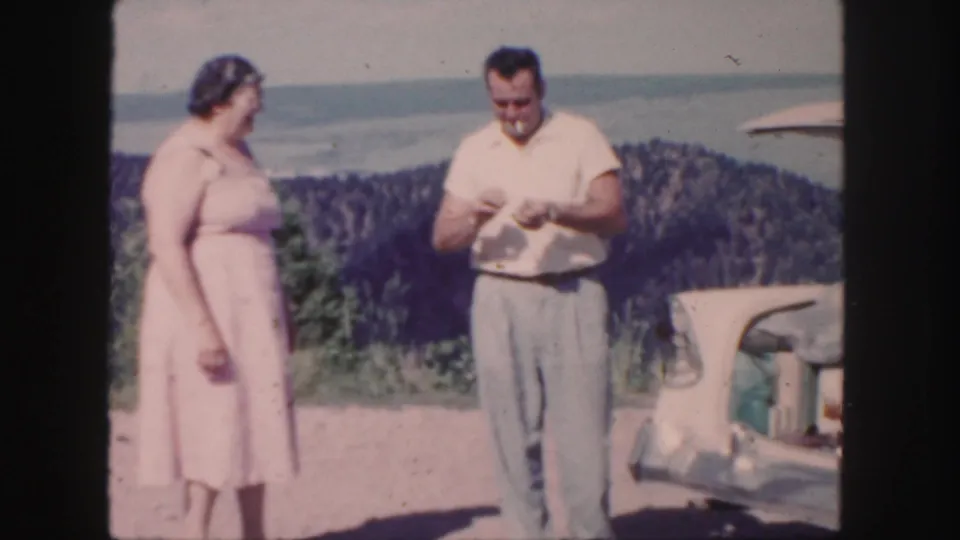 A woman and man stand by a classic car in 1958 Colorado home-movie footage, with the man looking down at an object in his hands.