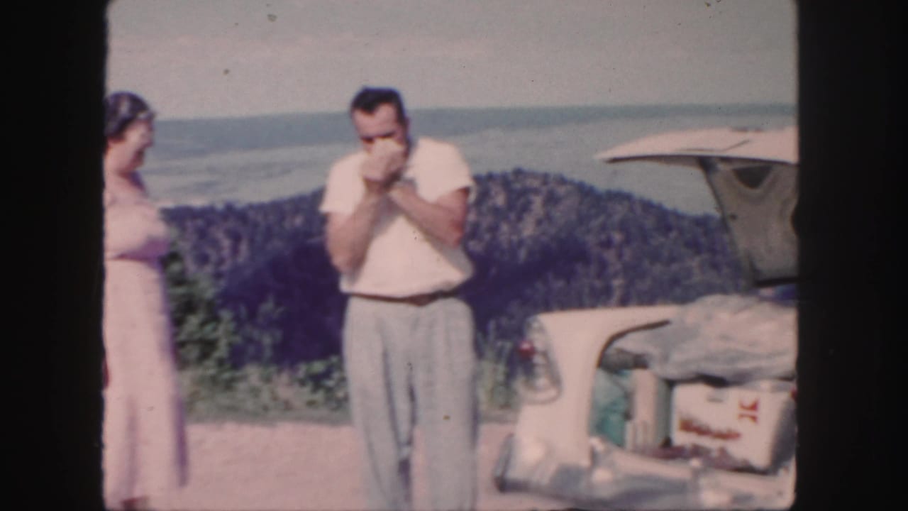 Man smoking beside an open car trunk while a woman stands nearby at a scenic overlook.