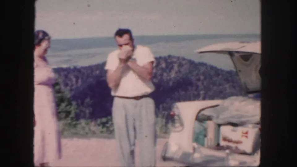 1958 Colorado home-movie frame showing a man with a cigarette beside an open car trunk as a woman in a pink dress stands nearby.