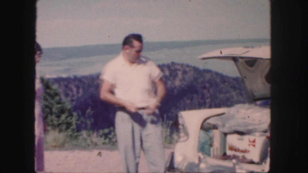 1958 Colorado frame with a man beside an open car trunk, the woman partly visible at left, and water and wooded hills behind them.
