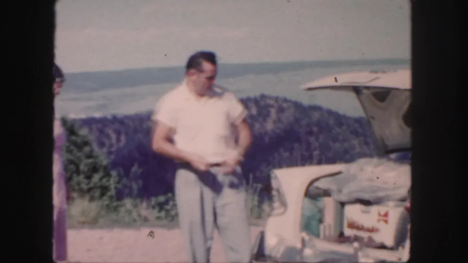 1958 Colorado frame with a man beside an open car trunk, the woman partly visible at left, and water and wooded hills behind them.
