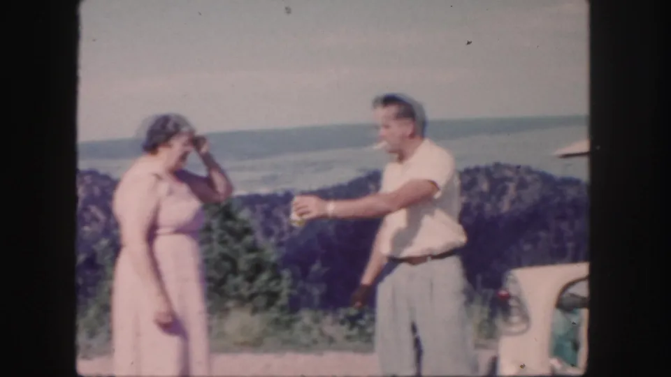 1958 Colorado home-movie frame showing the woman in a pink dress and the man gesturing near the open car trunk at a water overlook.