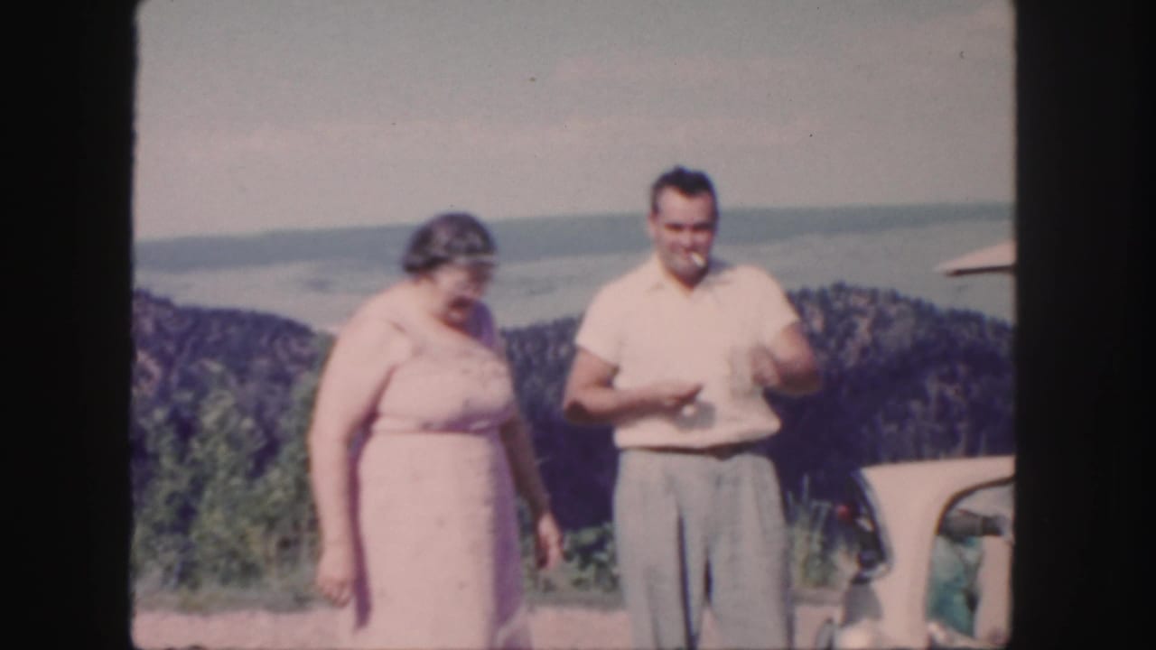 In 1958 Colorado home-movie footage, a woman in a pink dress and a man in a light shirt stand at a scenic mountain overlook.