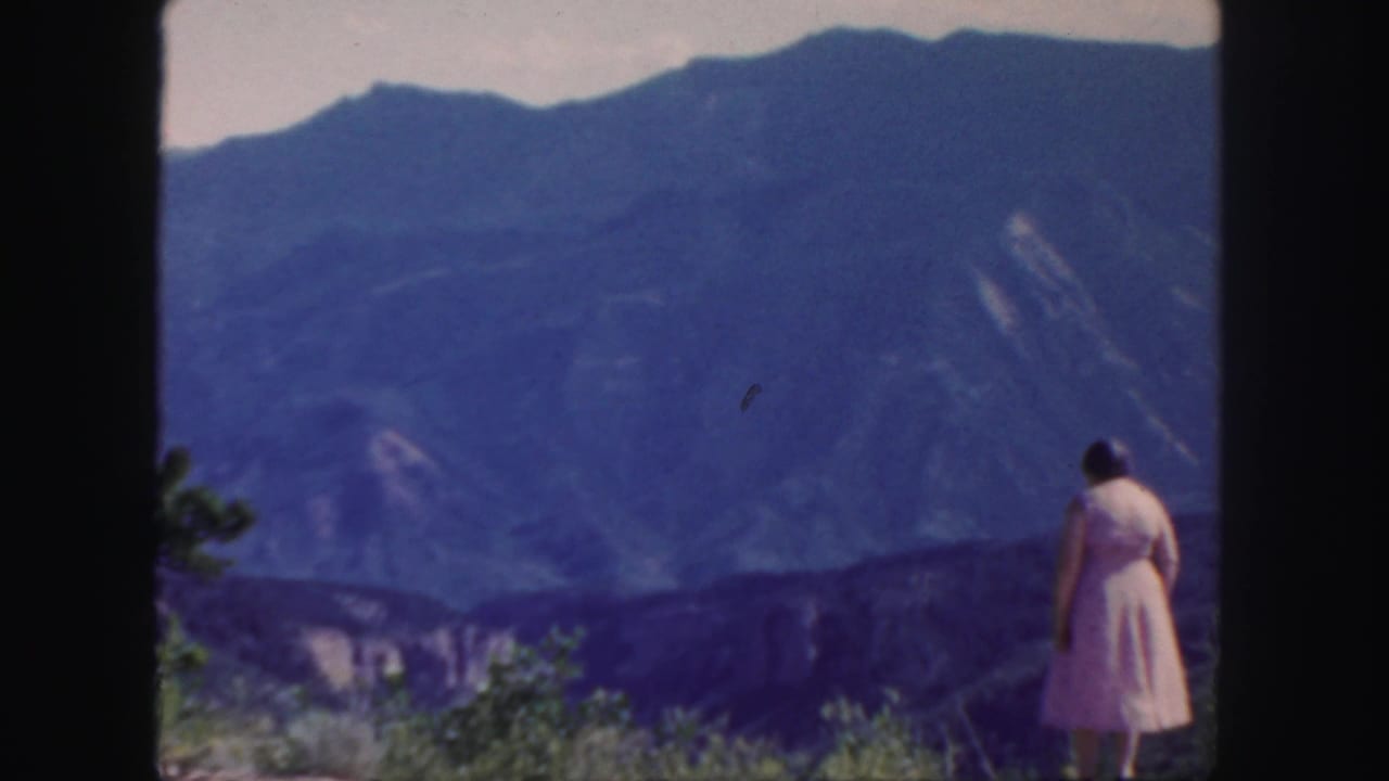 In 1958 Colorado footage, a woman in a pink dress stands on a hillside overlook facing distant blue mountains.