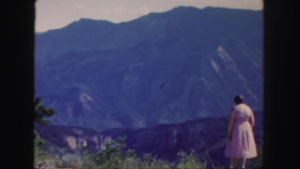 In 1958 Colorado footage, a woman in a pink dress stands on a hillside overlook facing distant blue mountains.