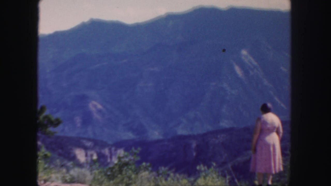 Woman stands at the right edge of a mountain overlook under a pale sky.