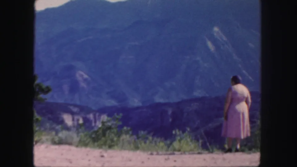 A distant woman in a pink dress stands on a Colorado hillside, framed by soft 8mm color and blue ridgelines.
