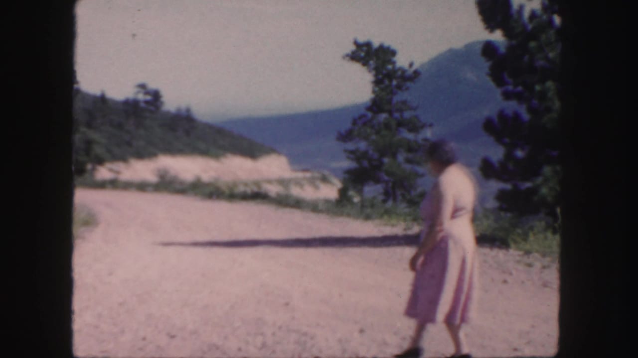 1958 Colorado home-movie frame of a woman in a pale dress walking along a sunlit dirt road below wooded mountain scenery.