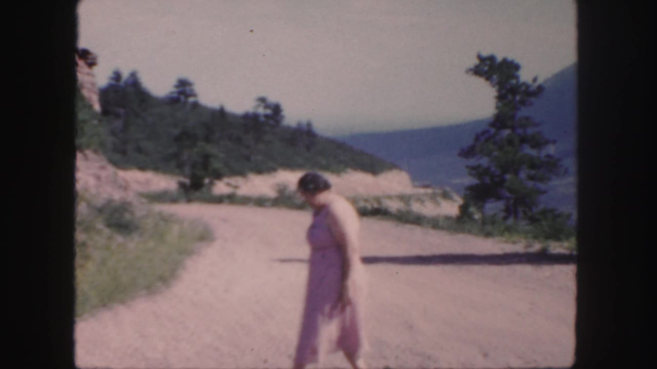Woman walking near the edge of a dirt road with a wooded slope and mountains beyond.
