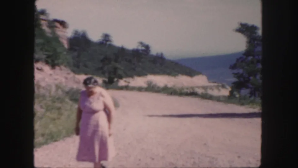 1958 Colorado home-movie frame with the woman nearer the camera on a dirt road, backed by grass, trees, and hazy mountain slopes.