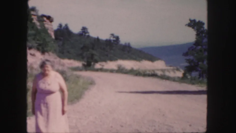 1958 Colorado frame of a woman in a pale dress near the foreground on a sunlit dirt road, with trees and mountain slopes beyond.
