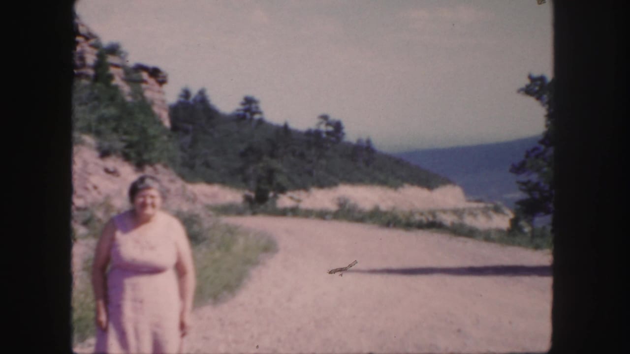 Older woman in a pale pink dress faces the camera on a winding Colorado mountain road in 1958 home-movie footage.