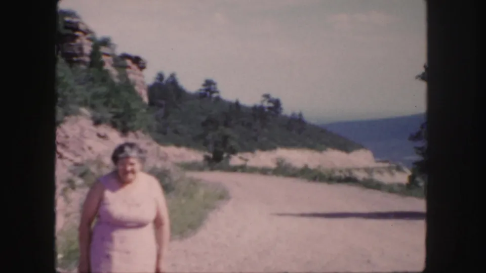 Older woman smiles toward the camera beside a dusty Colorado road, with pines, rocks, and distant hills visible.