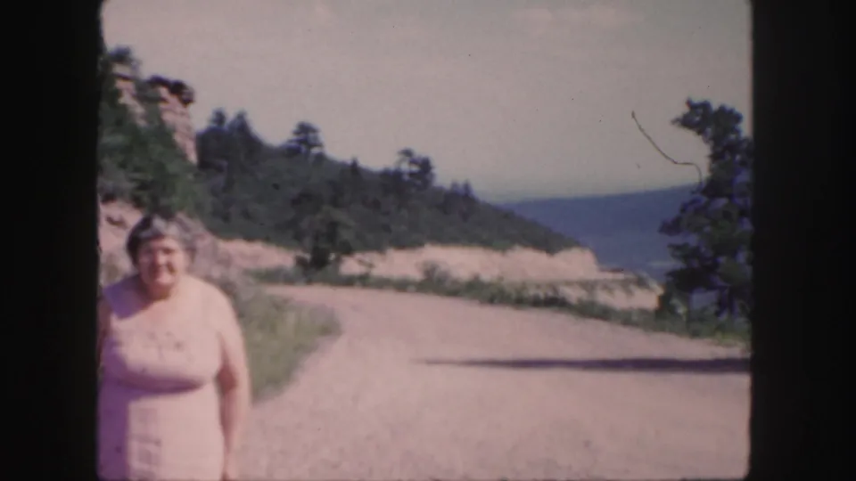 A handheld 1958 Colorado frame places the older woman near the edge while the mountain road and rocky slope fill the view.