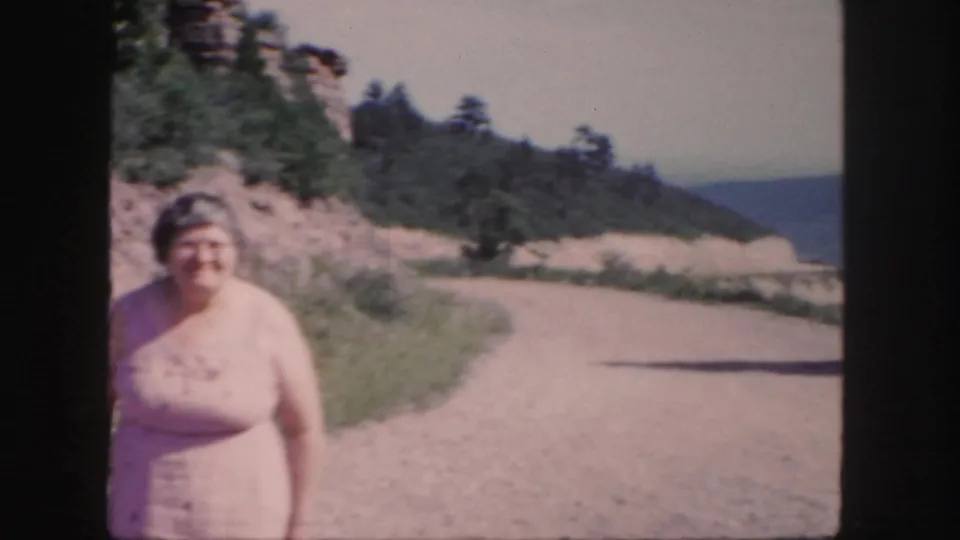 Older woman in a pale dress appears closer to the camera on the Colorado roadside, smiling against a dry mountain backdrop.