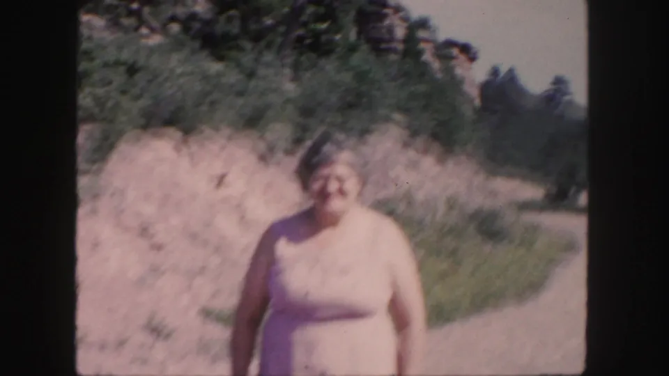 Soft 8mm Colorado frame of a woman in a pale dress standing along a curving dirt path in bright daylight.
