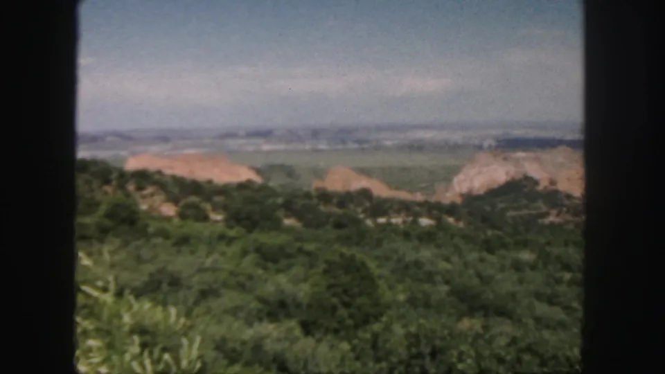 1958 Colorado landscape view showing green foreground hills, tan rock formations, a broad valley, and pale sky.