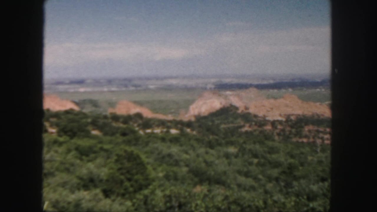 Soft 8mm frame looking over green hills, reddish rock formations, distant ridges, and pale sky.