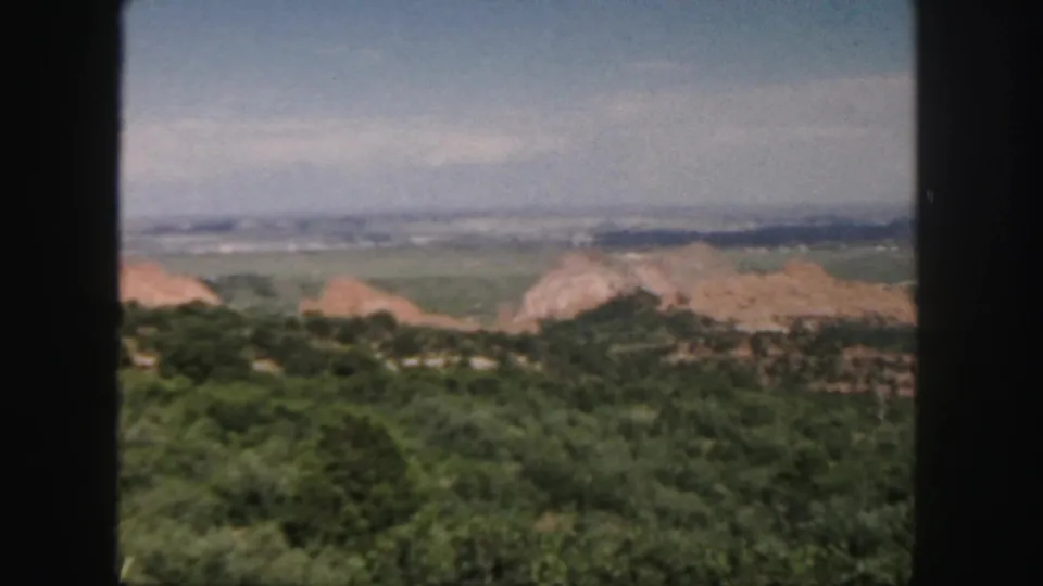 1958 Colorado home-movie frame showing a high overlook with green foreground, reddish rock formations, distant ridges, and pale blue sky.