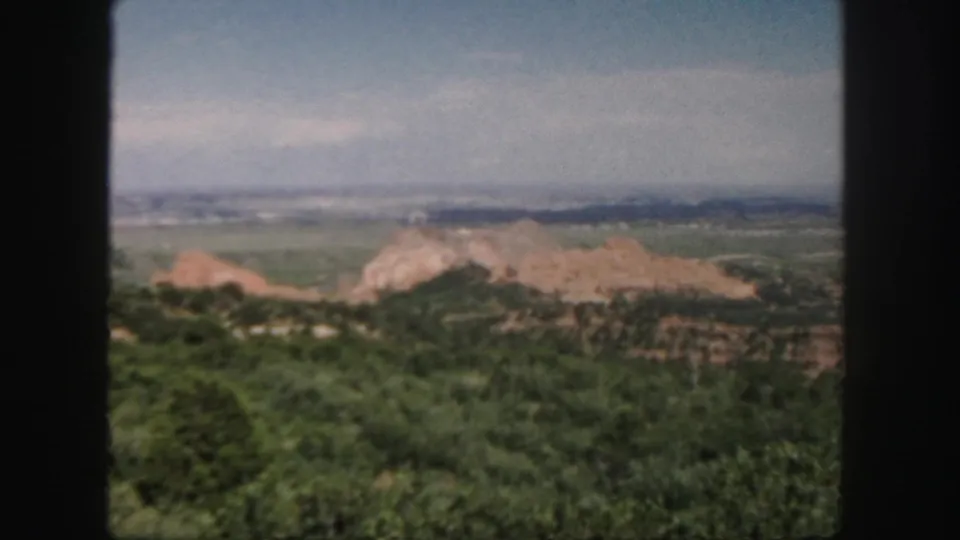1958 Colorado home-movie view from a high vantage point, with green terrain, reddish outcrops, distant ridges, and a blue-gray sky.